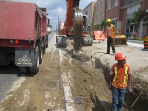 Excavator digging a trench beside a dump truck while workers look on
